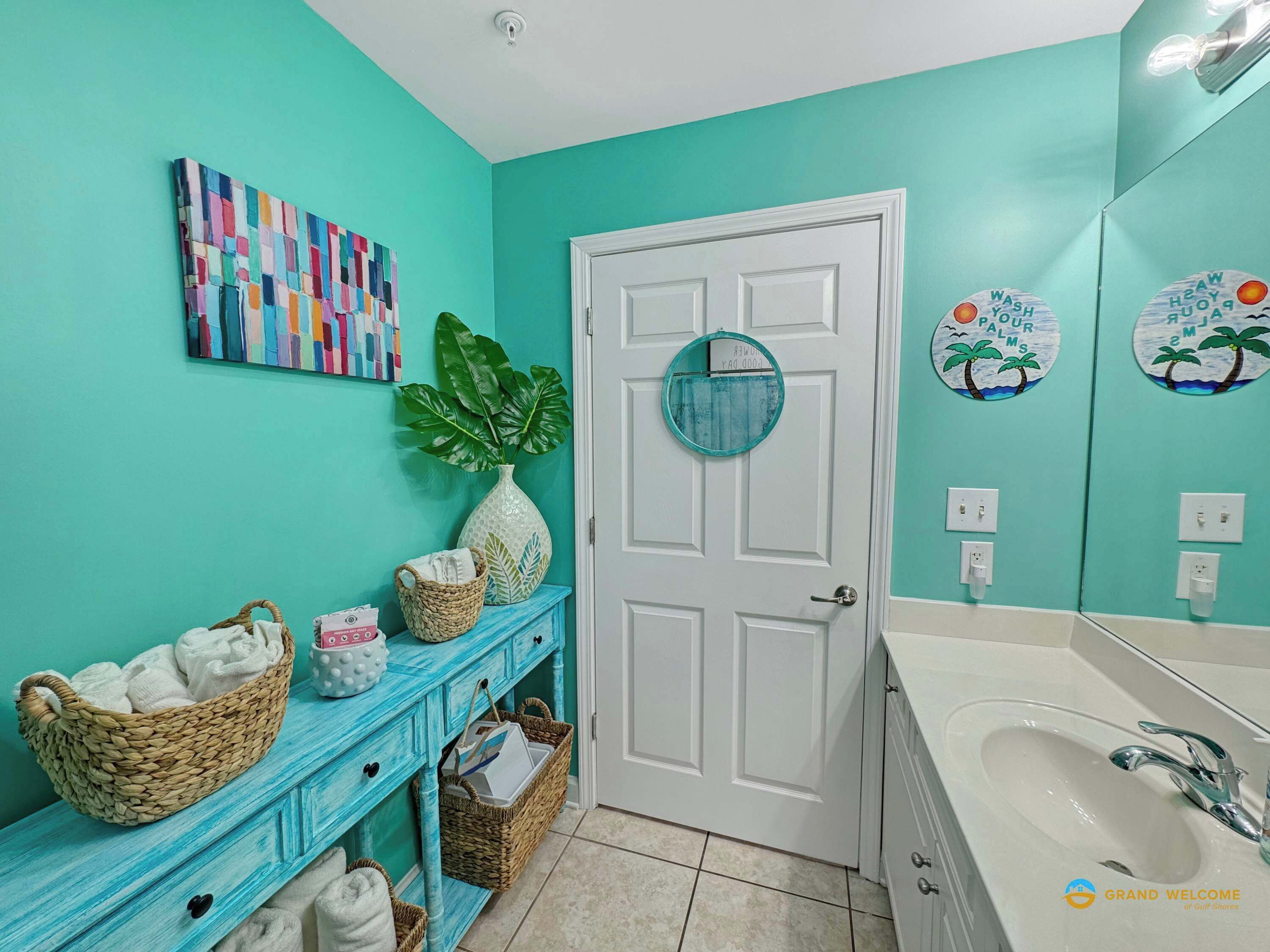 Une salle de bain aux murs de couleur turquoise, avec une porte blanche, un grand miroir et un lavabo doté d’un robinet chromé. On y trouve des paniers sur un meuble-lavabo bleu et une plante accrochée au mur.
