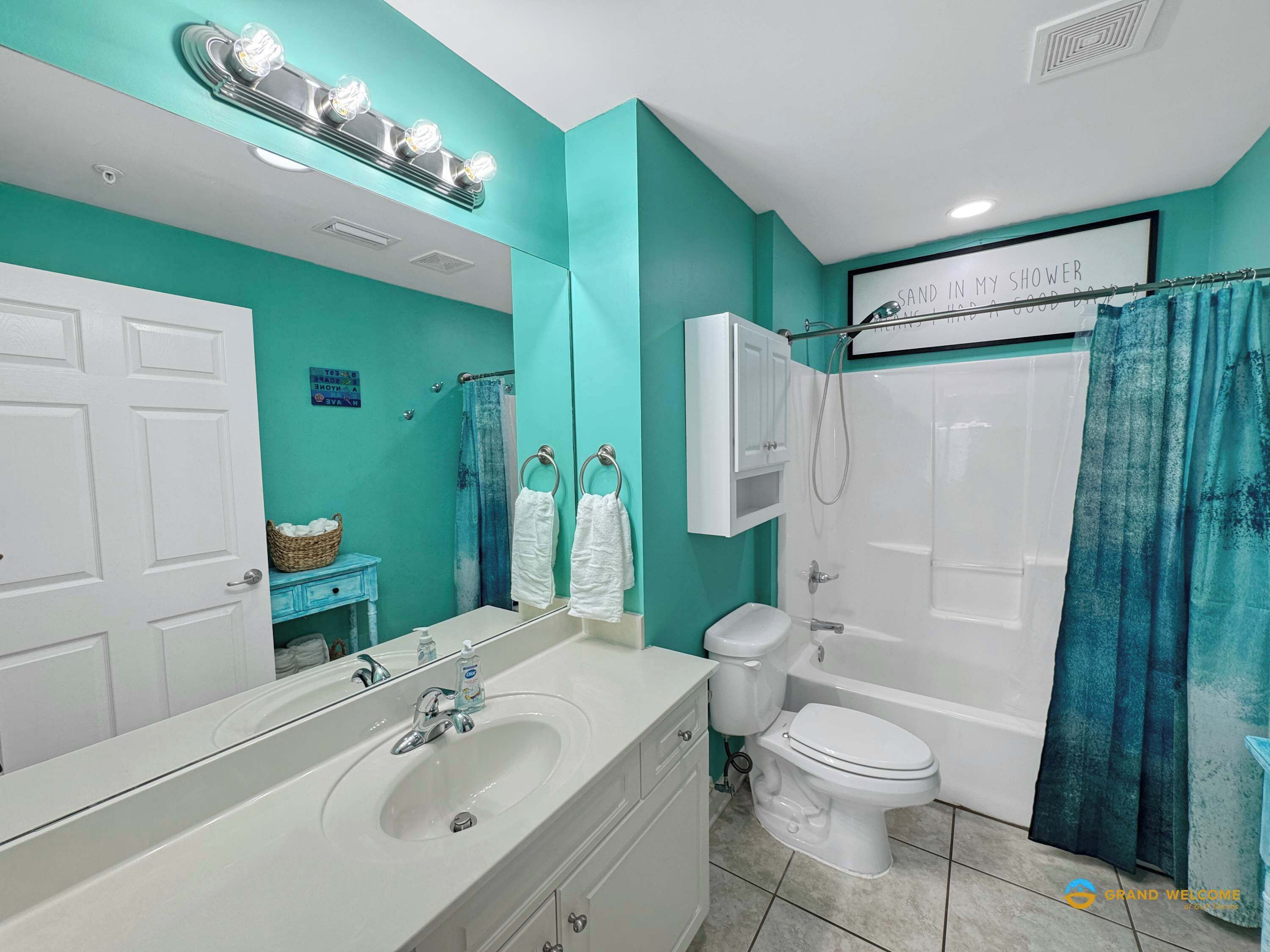 Une salle de bain avec un lavabo, des toilettes et une baignoire, tous de couleur blanche. Un miroir avec des lumières est placé au-dessus du lavabo, et une pancarte indique : « SABLÉ DANS MA DOUCHE ».