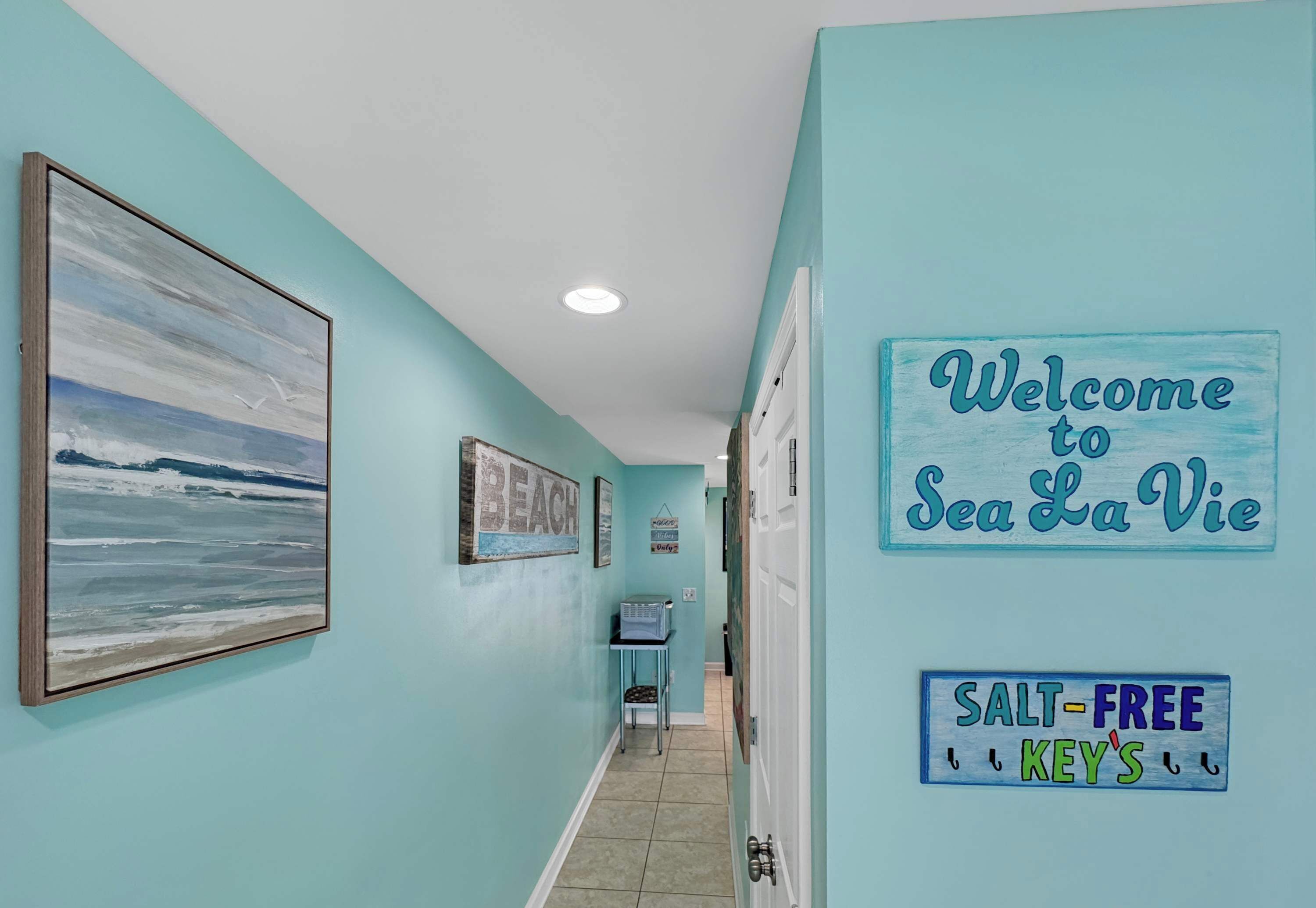 Ein Flur mit einem 'Welcome to Sea La Vie' Schild, einem 'Salt-Free Keys' Schild und einem Strandgemälde.
