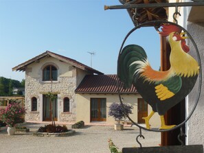 A stone building with a tiled roof, a rooster sign, and a garden with flowers and a tree. - Grange Carrée (Saint-Rémy)