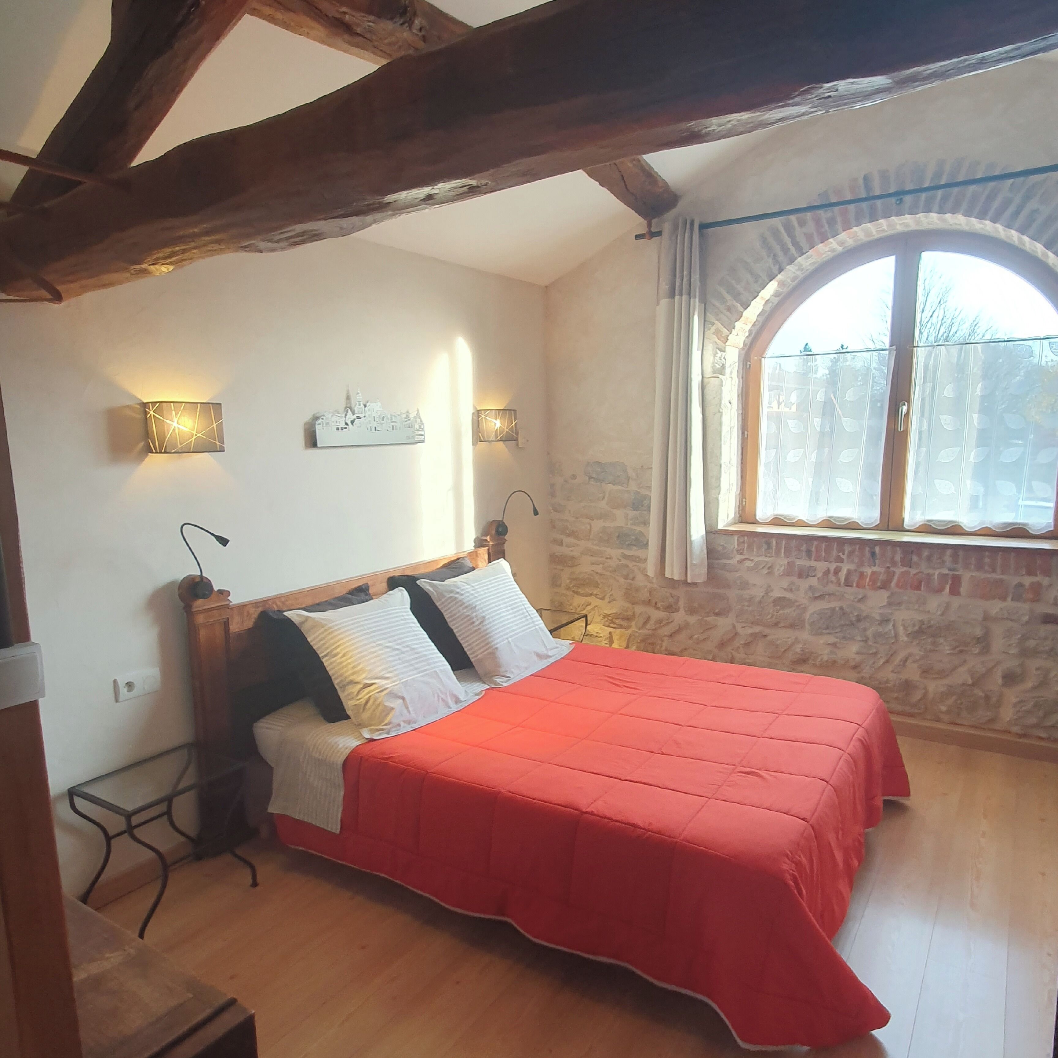 A bedroom with a wooden bed frame, a red bedspread, a stone wall, and a window with a view of trees.