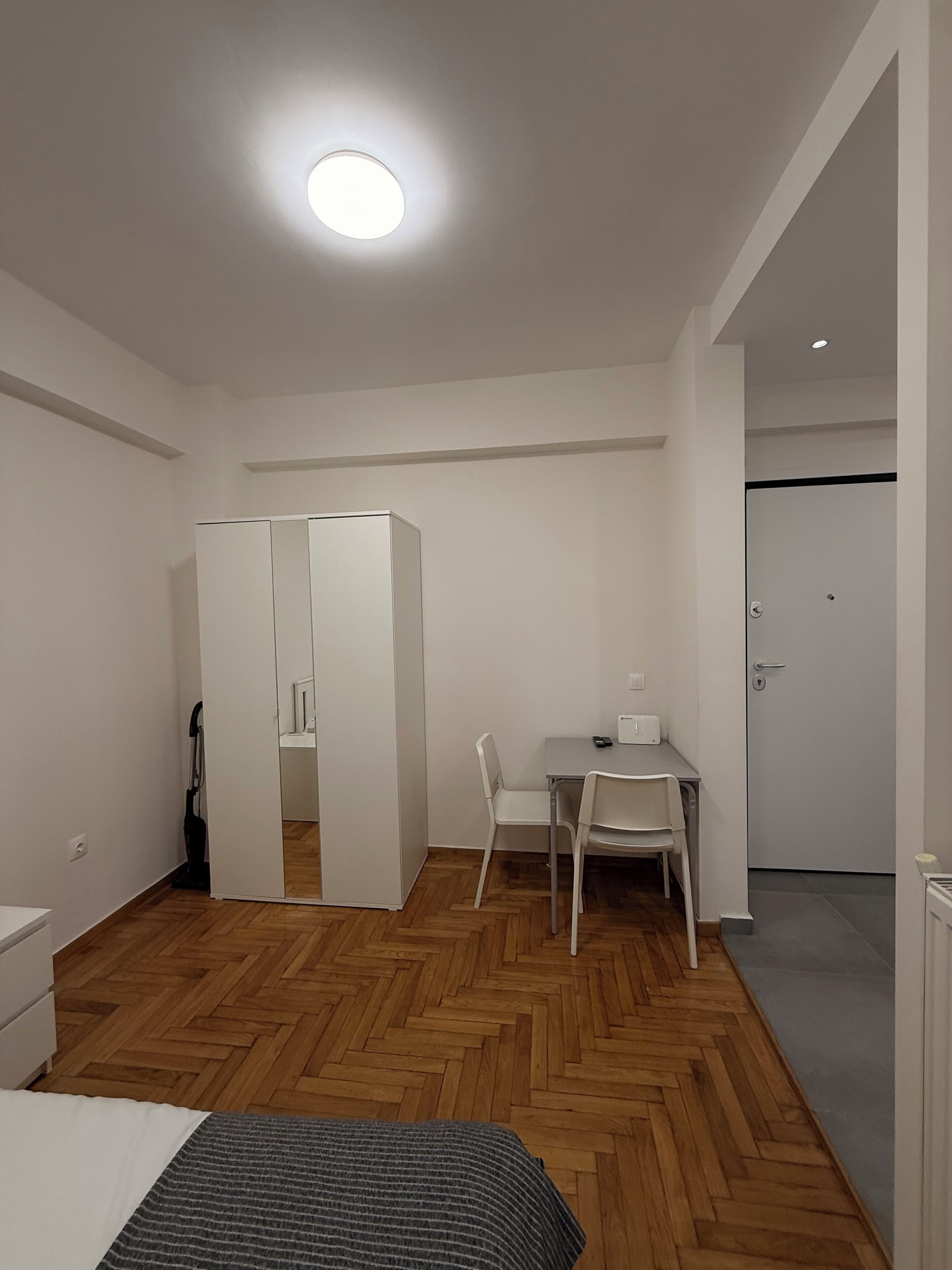 A modern, minimalist room with a wooden herringbone floor, a bed, a desk with two chairs, a wardrobe, and a door.