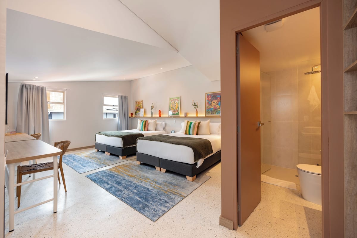 Quarto de hotel moderno com duas camas, área de refeições e chuveiro.