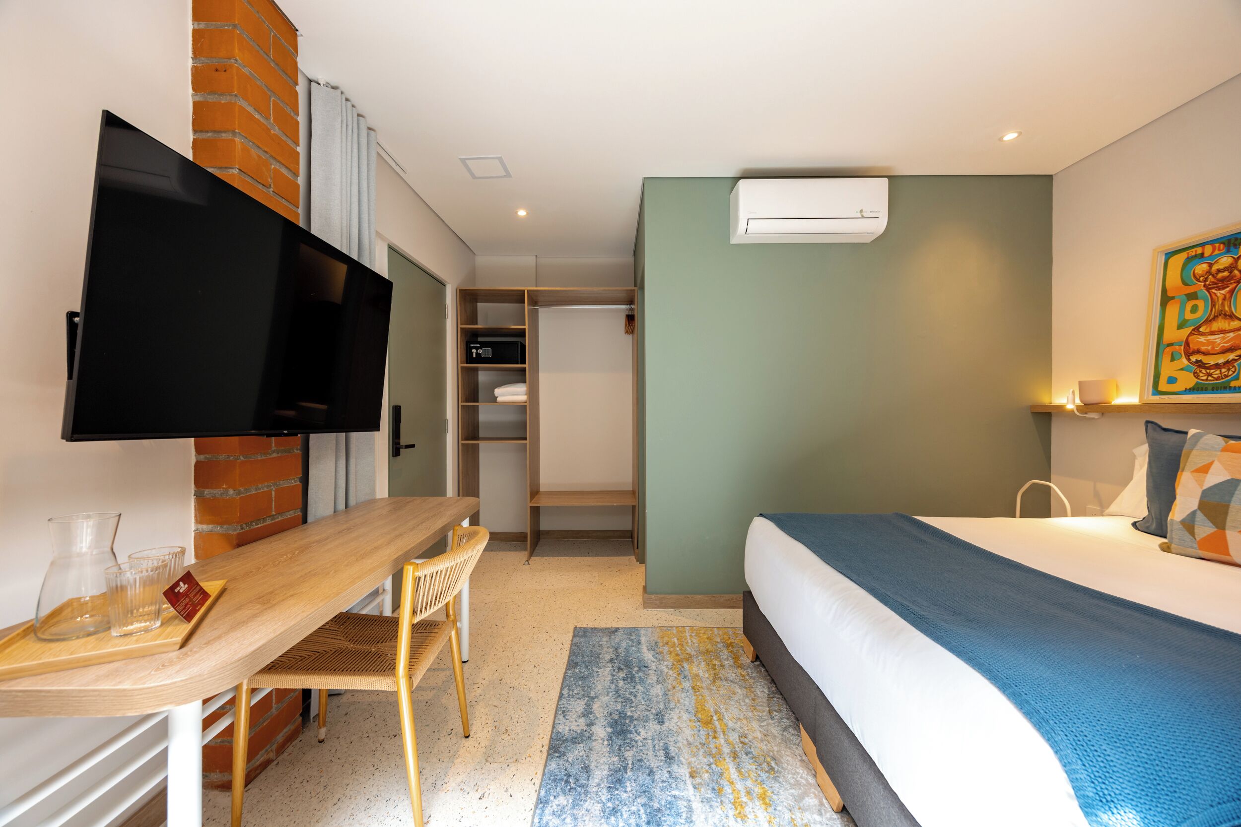 A hotel room with a bed, a desk with a chair, a television, a wardrobe, and an air conditioning unit.