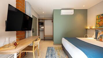 A hotel room with a bed, a desk with a chair, a television, a wardrobe, and an air conditioning unit.