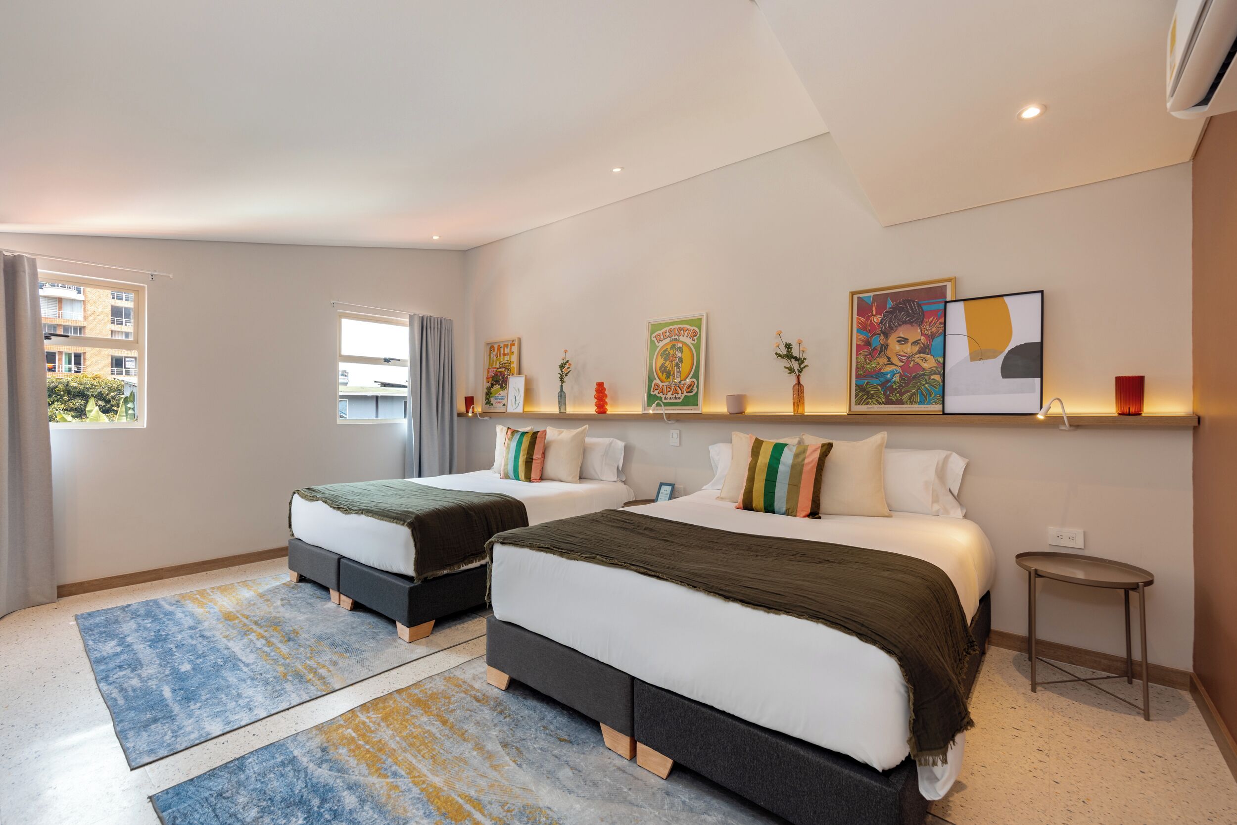 A modern hotel room with two beds, a small table, a window, and artwork on the wall.