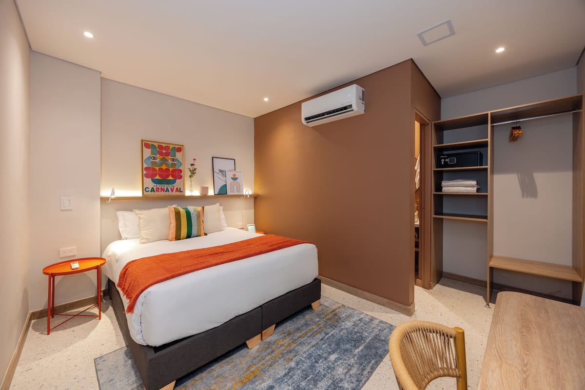 Quarto de hotel com cama, uma mesinha, uma cadeira, guarda-roupa e ar condicionado.