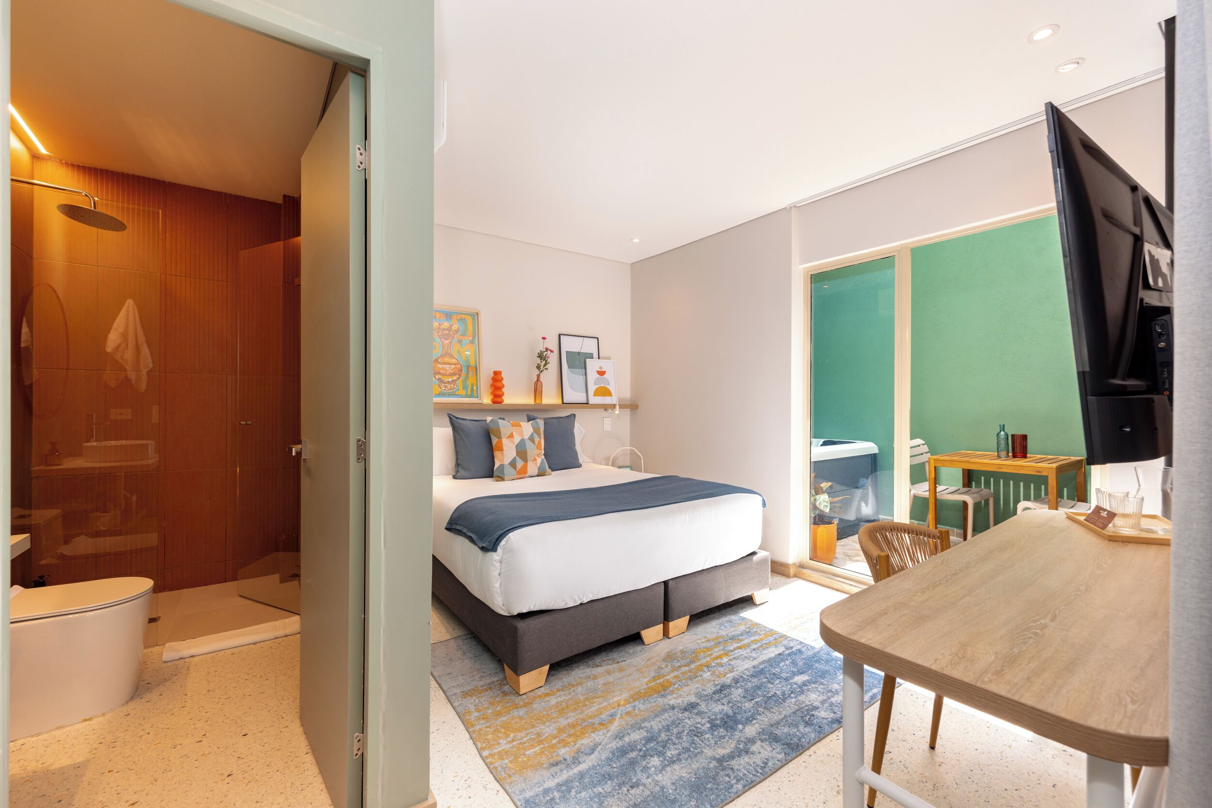 A modern hotel room with a bed, a bathroom, a dining area, and a TV.