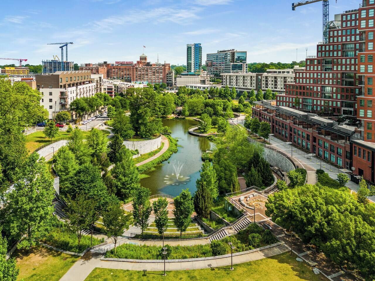 A park with a pond, surrounded by trees and pathways, with buildings in the background.