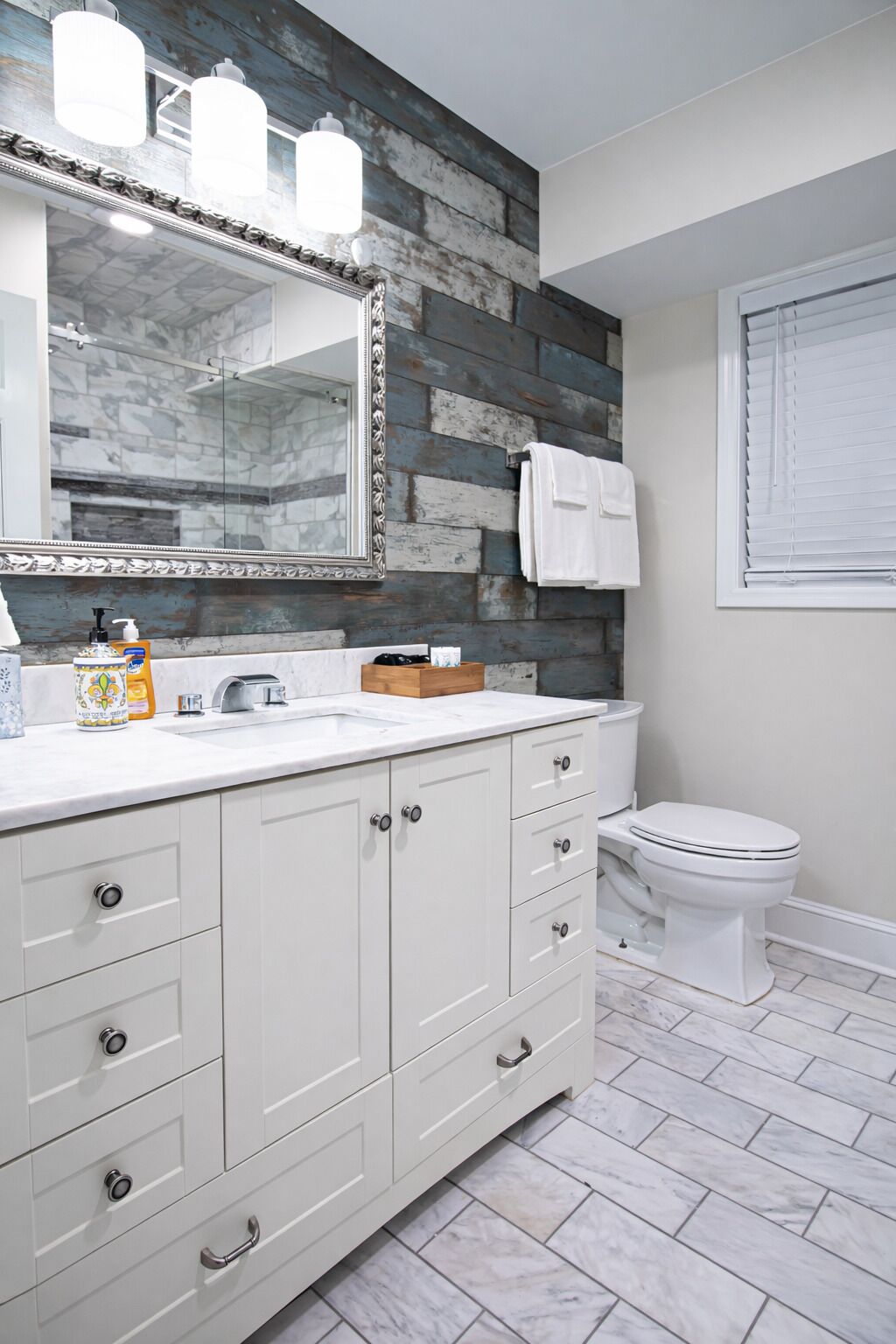 Une salle de bain avec un meuble-lavabo blanc, un miroir, un mur d’accent en bois, des toilettes et une fenêtre avec des stores.