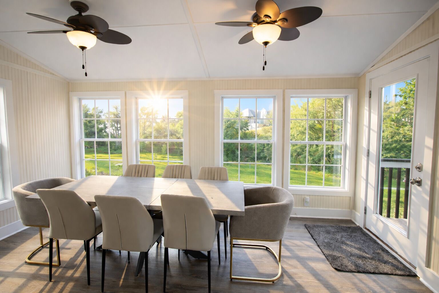 Une véranda dotée d’une grande table et de chaises, de deux ventilateurs de plafond et de grandes fenêtres offrant une vue sur un paysage verdoyant.