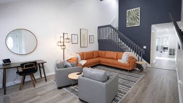 Un salon moderne avec un canapé d’angle, un miroir rond, un bureau et un escalier.