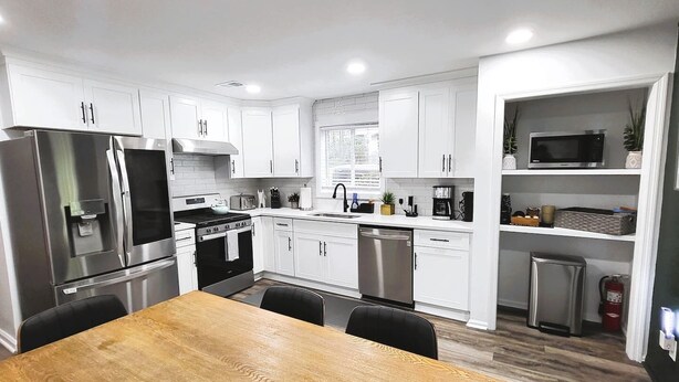 A modern kitchen with white cabinets, stainless steel appliances, a wooden dining table, and a built-in microwave shelf. - 7 BR / 5 BA | 2 Homes Sandy Springs Near Buckhead (Atlanta)