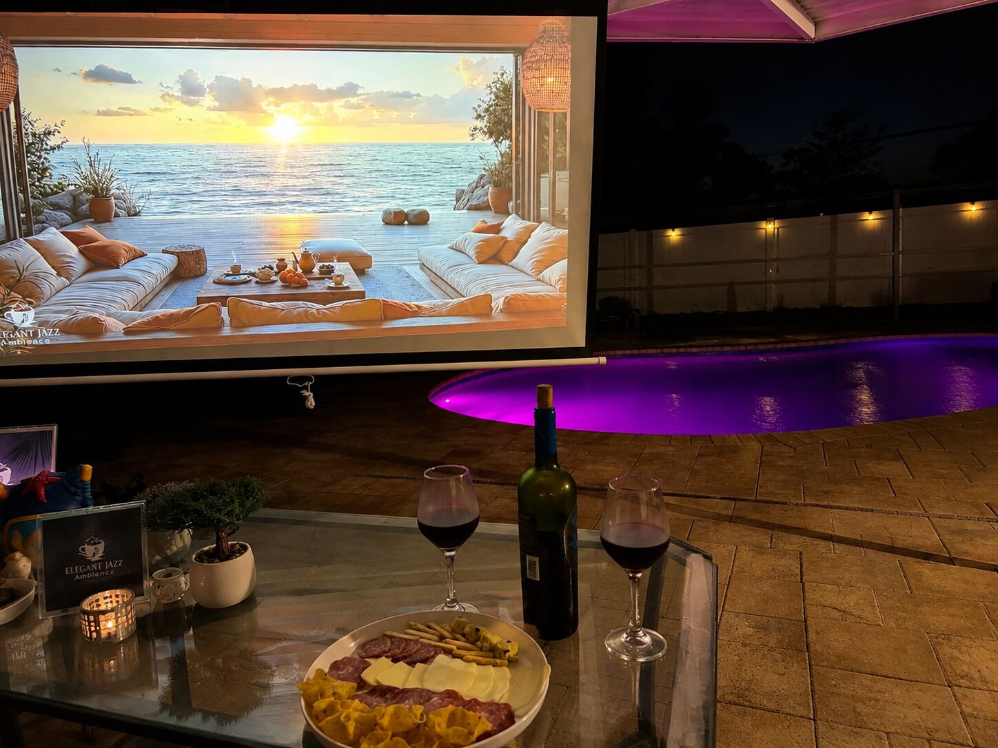 A poolside dining area with a sunset view, a dining table with food and drinks, and a large screen displaying a relaxing scene.