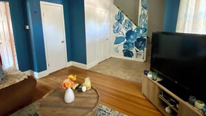 Un salon avec un mur bleu, une table basse en bois ornée de fleurs, un meuble pour la télévision et un escalier dont les murs sont recouverts d’un papier peint à motifs floraux.