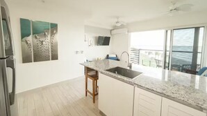Una cucina moderna con un piano di lavoro in granito, un lavello e uno sgabello in legno. Una grande finestra offre vista sull'oceano.