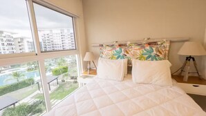 Camera da letto con un letto grande, due lampade ai lati del letto e vista su una piscina e degli edifici.