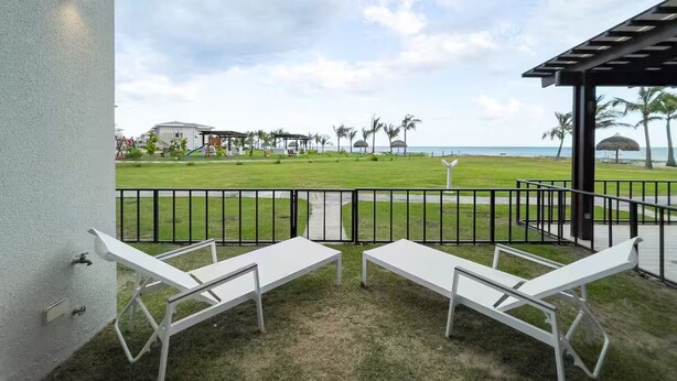 Uma varanda com duas cadeiras de descanso brancas, vista para uma área gramada com palmeiras e uma praia ao longe.