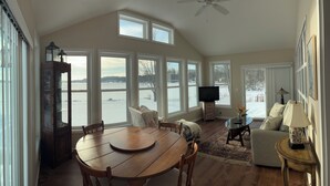 A spacious living room with a large wooden dining table, a television, and a cozy seating area with a view of a lake. - Lakefront 2-bedroom home with dock and AC, minutes from Chautauqua, Sleeps 6 (Mayville)