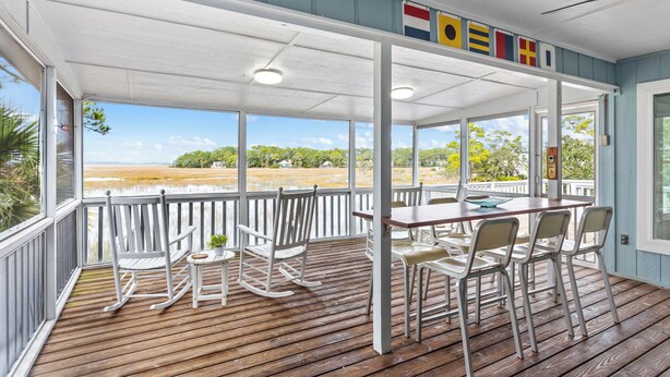 A covered outdoor area with a wooden deck, white rocking chairs, and a table with chairs. - Remodeled Center Island Home, Marsh Views, 2 Golf Carts, Stellar Sunsets (Fripp Island)