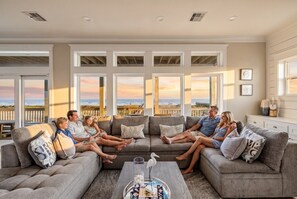 A living room with a sectional sofa, a coffee table with books, and large windows with a view. - Beachfront Home- Hot Tub & Private Walkover (Galveston)