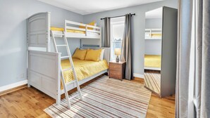 Habitación con literas, suelo de madera, estructura de cama blanca, colcha amarilla, mesita de noche pequeña con lámpara y ventana con cortinas.