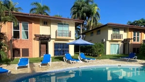 A pool area with blue lounge chairs and umbrellas, surrounded by a building with a tiled roof and windows. - Villa Postosomar Cabarete (Cabarete)