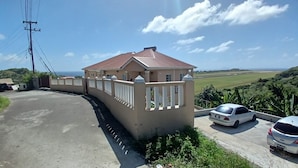 Una casa grande con un camino de entrada cerrado por una reja, un coche blanco estacionado al frente y vistas a un paisaje verde.