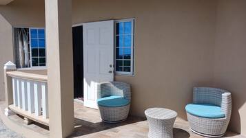 A covered outdoor seating area with wicker chairs and a small table, a white door, and a window with a view.