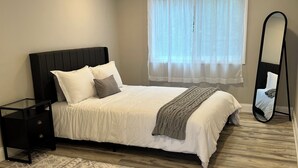 Un dormitorio con cama, mesita de noche, espejo y ventana con cortinas traslúcidas.