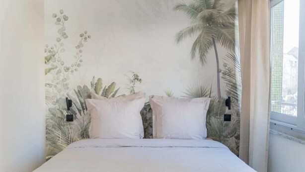 Une chambre à coucher avec un lit, deux oreillers, une fresque murale représentant des plantes et une fenêtre avec des rideaux.