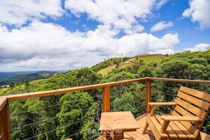 Eine Holzterrasse mit einer Bank, die auf eine üppige, grüne Landschaft mit Hügeln und blauem Himmel blickt.