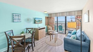 Ein Zimmer mit Esstisch, Stühlen, einer Couch und einem Fernseher. Durch große Fenster ist der Blick aufs Meer möglich.
