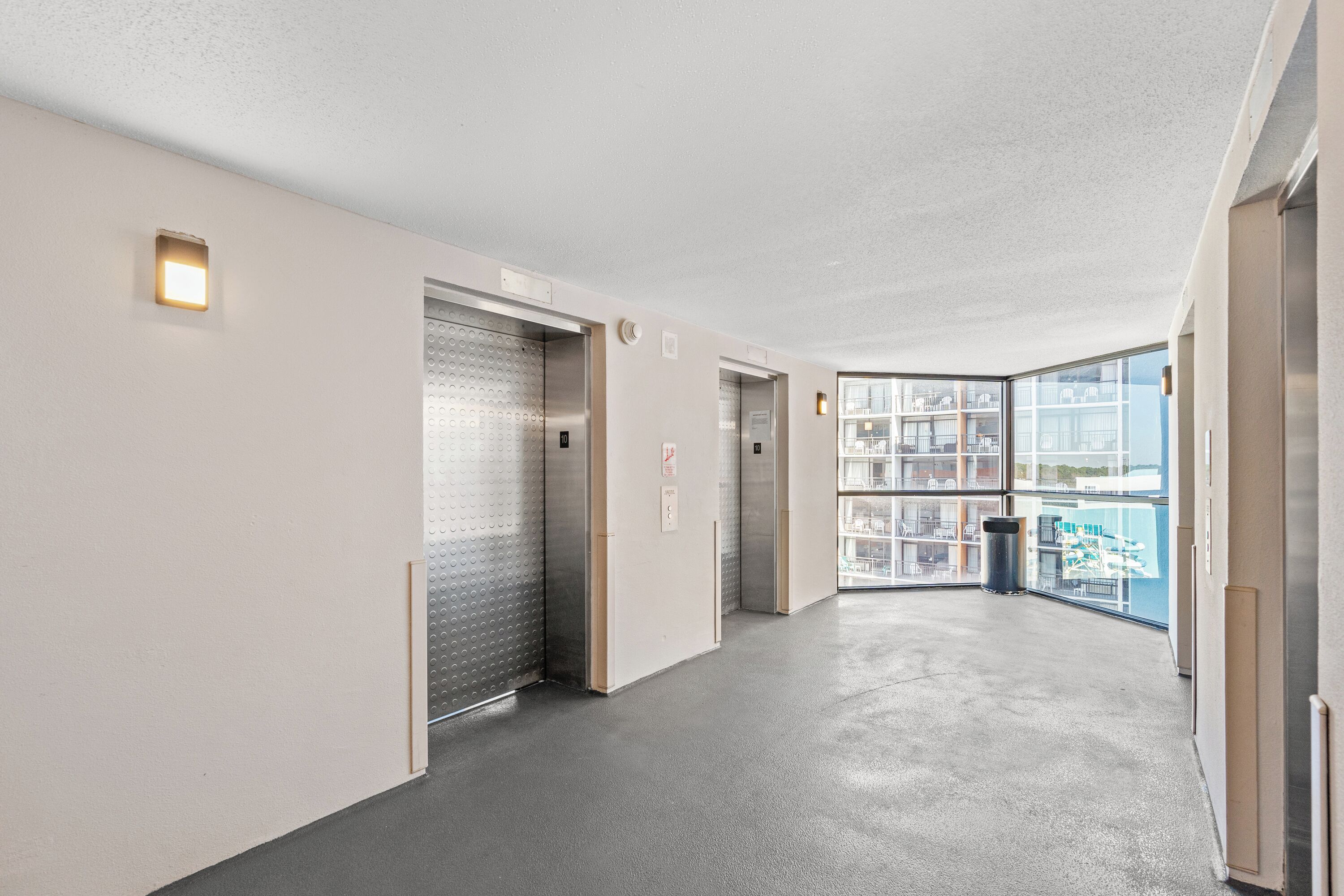 A modern hallway with elevators, a trash bin, and large windows offering a view of the city.