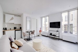 Una sala de estar moderna con un sofá, un reposapiés redondo, un comedor con sillas, una cocina con refrigerador y un televisor montado en la pared.