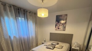 Ein Schlafzimmer mit Bett, Nachttischen, einer Lampe, Deckenleuchte, einem Fenster mit Vorhängen und einem Wandbild.