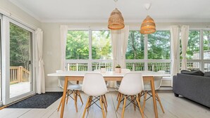 Un espace repas moderne avec une table et des chaises blanches, deux lampes suspendues en osier et de grandes fenêtres donnant sur un paysage verdoyant.