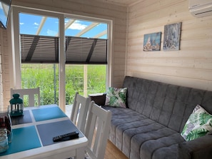 Ett rum med en grå soffa, ett bord med en fjärrkontroll och ett fönster med utsikt över grön gräsmatta.