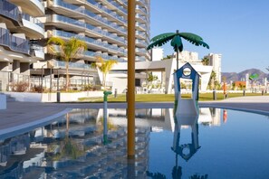 Un bâtiment moderne doté de balcons, d’un bassin aux reflets changeants et d’une sculpture représentant un palmier.
