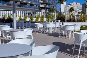 Terrasse avec des tables rondes et des chaises blanches, un bâtiment moderne doté de balcons, et une vue dégagée sur les palmiers et les autres bâtiments en arrière-plan.