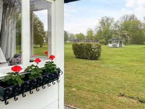 A view from a window with a white exterior, a black metal planter with red flowers, and a well-maintained lawn with a gazebo and a hedge. - 6 Person Holiday Home in Lottorp (Löttorp)