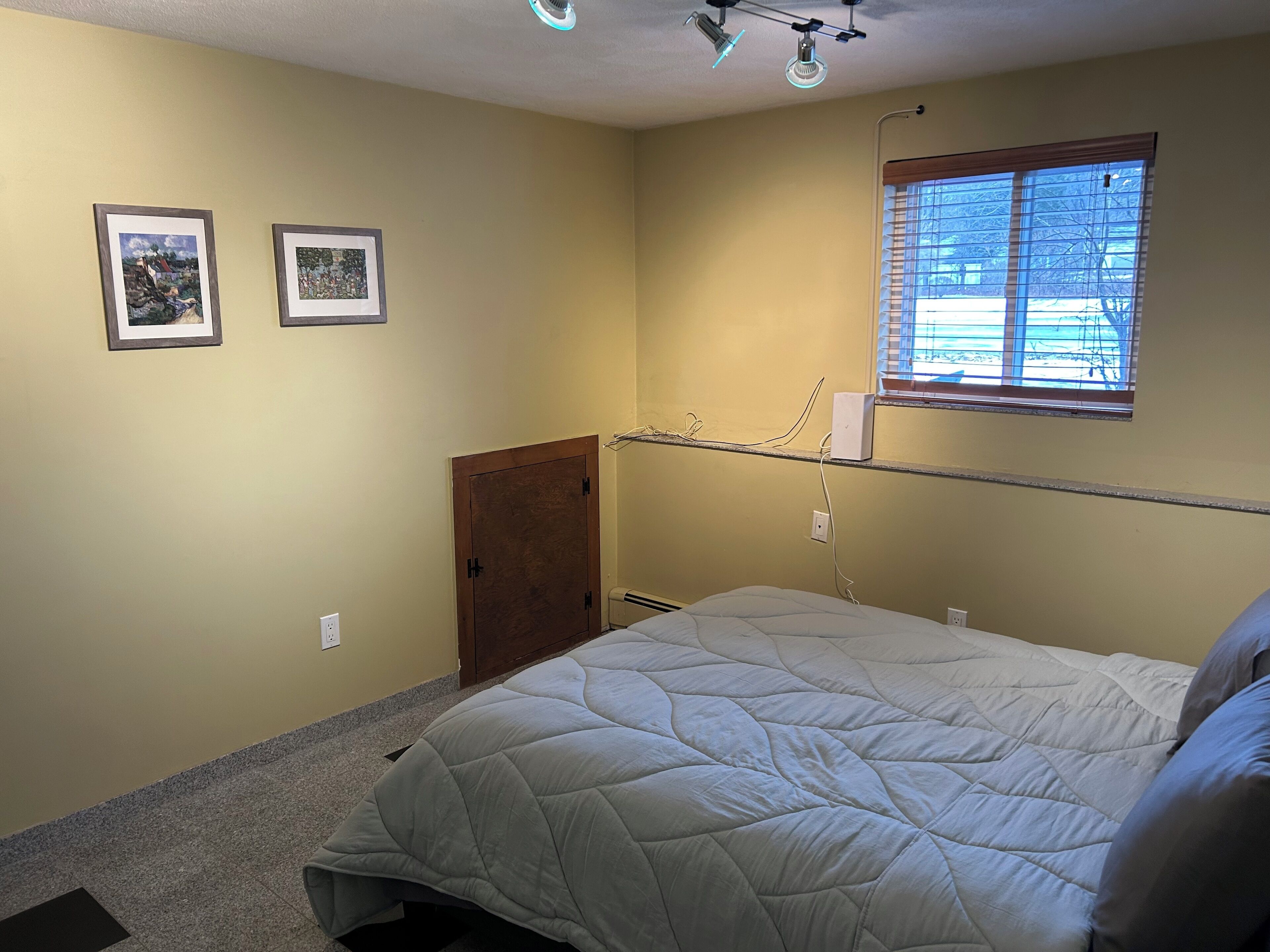 Ein Schlafzimmer mit einem Bett, zwei gerahmten Bildern an der Wand, einem Fenster mit Jalousien und einer Tür.