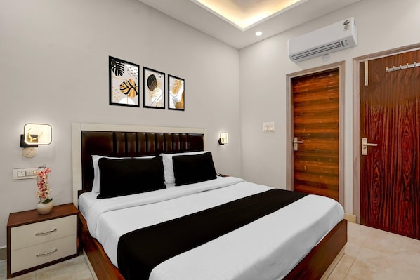 A hotel room with a bed, bedside table, wall-mounted air conditioning unit, and a wooden door.