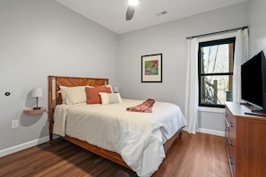 A bedroom with a wooden bed, white bedding, a bedside lamp, a TV on a wooden cabinet, and a window with curtains.