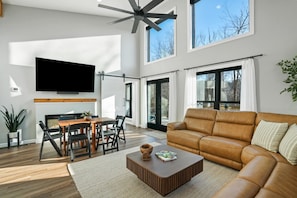 A modern living room with a leather sofa, a flat-screen TV mounted on the wall, a dining area with a wooden table and chairs, and large windows allowing natural light.