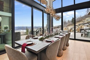 Ein Essbereich mit einem langen Tisch, gedeckt für sechs Personen, großen Fenstern mit einem schönen Ausblick und einer modernen Pendelleuchte.