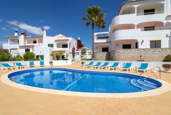 A swimming pool with lounge chairs, surrounded by white buildings with balconies and a stone wall. - Casa Franjolea - 2 Bedrooms,  Shared Pool & Ocean View (Carvoeiro)
