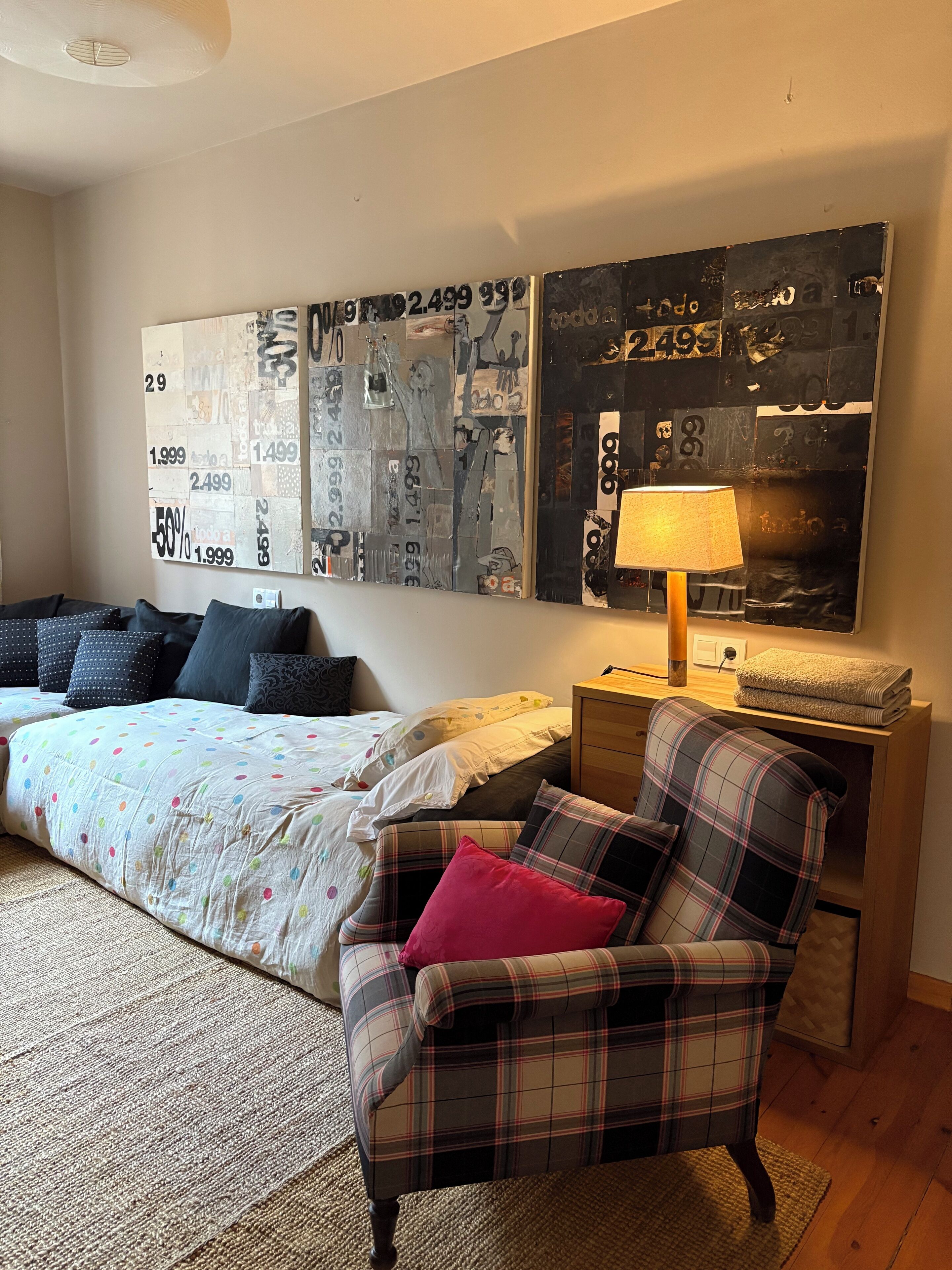 Een slaapkamer met een bed, een geruit fauteuil, een bijzettafeltje met een lamp en een groot abstract kunstwerk aan de muur.