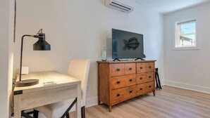 Una habitació amb escriptori de fusta, una cadira, un televisor sobre un moble de fusta i un aparell d'aire condicionat a la paret.