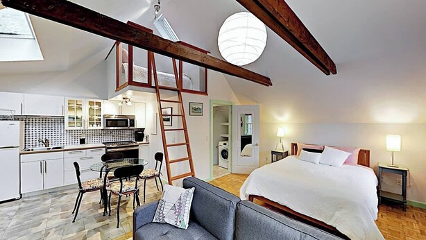 Ein Schlafzimmer im Loft-Stil mit einem großen Bett, einem grauen Sofa und einem Essbereich mit Tisch und Stühlen. Eine Leiter lehnt an der Wand, und ein Oberlicht ist in der Decke sichtbar.